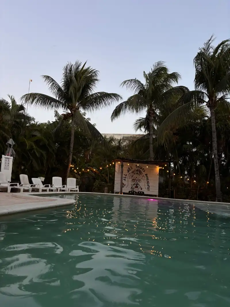 La alberca se ve al atardecer, con tumbonas blancas, palmeras alrededor y un mural iluminado con una guirnalda de luces.