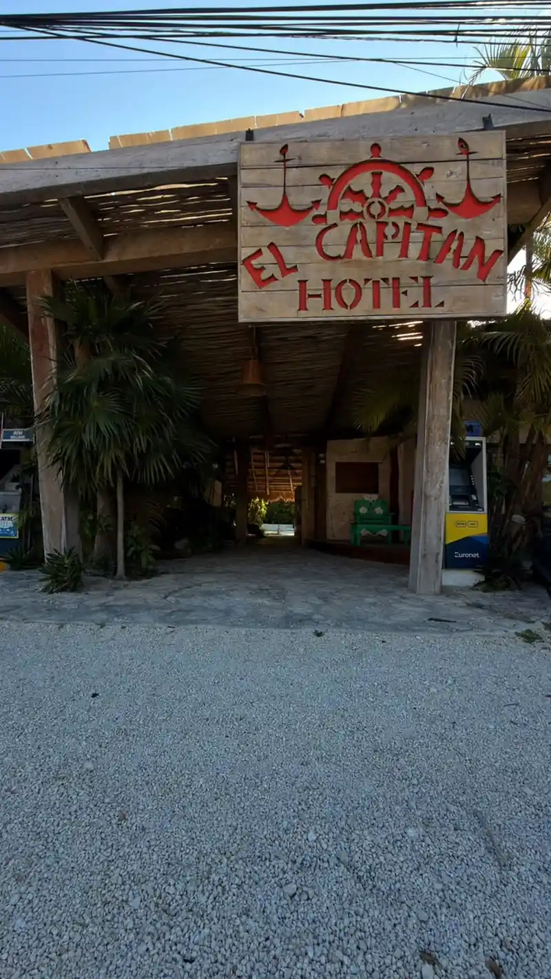 La entrada del hotel queda bajo una palapa de madera, con el rótulo tallado 'EL CAPITAN HOTEL' en rojo y blanco, piso de grava y un cajero automático junto a la columna derecha.