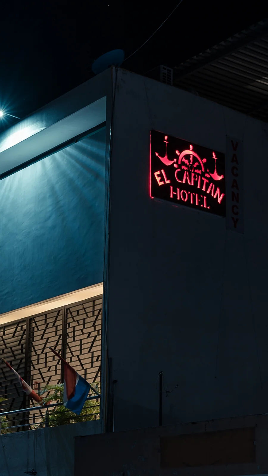 La fachada del hotel de noche: un letrero de neón rojo con el ancla y el timón de 'EL CAPITAN HOTEL' sobre el muro, el piso superior iluminado y banderas de varios países en la terraza.