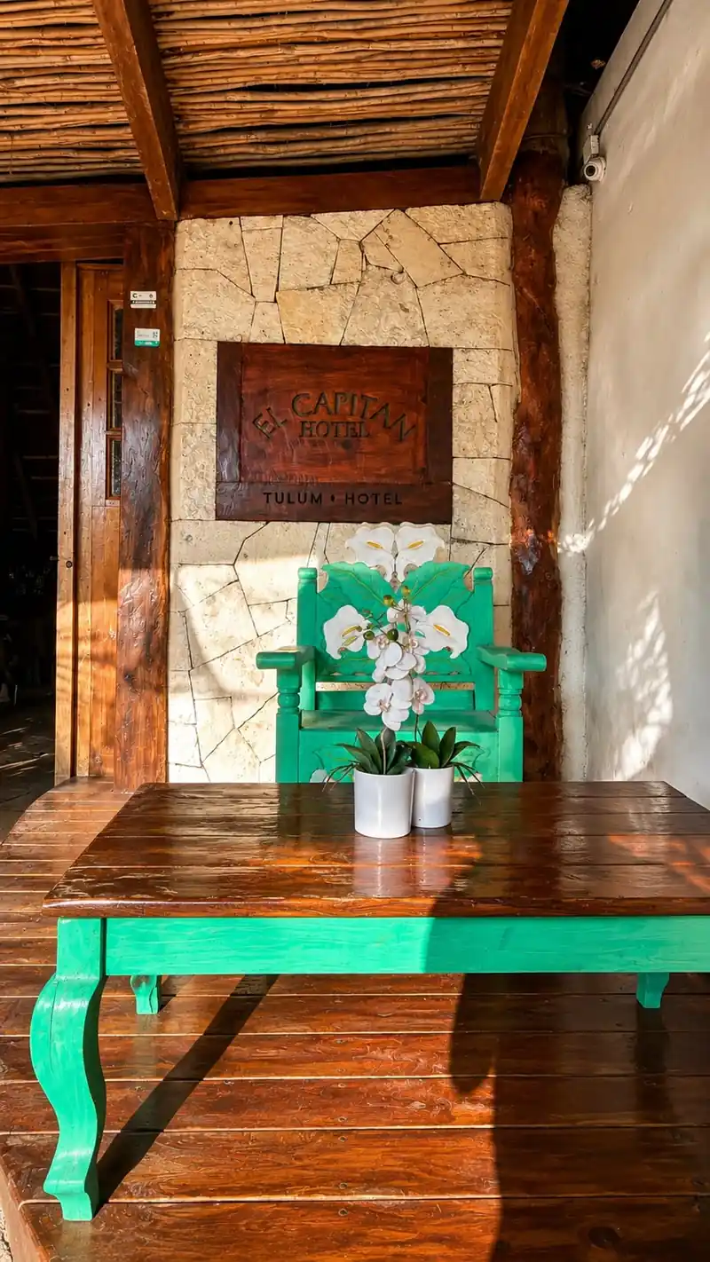 Una placa de madera con el texto 'EL CAPITAN HOTEL · TULUM · HOTEL' está empotrada en un muro de piedra, junto a una silla y una mesa verdes talladas con flores blancas en macetas.